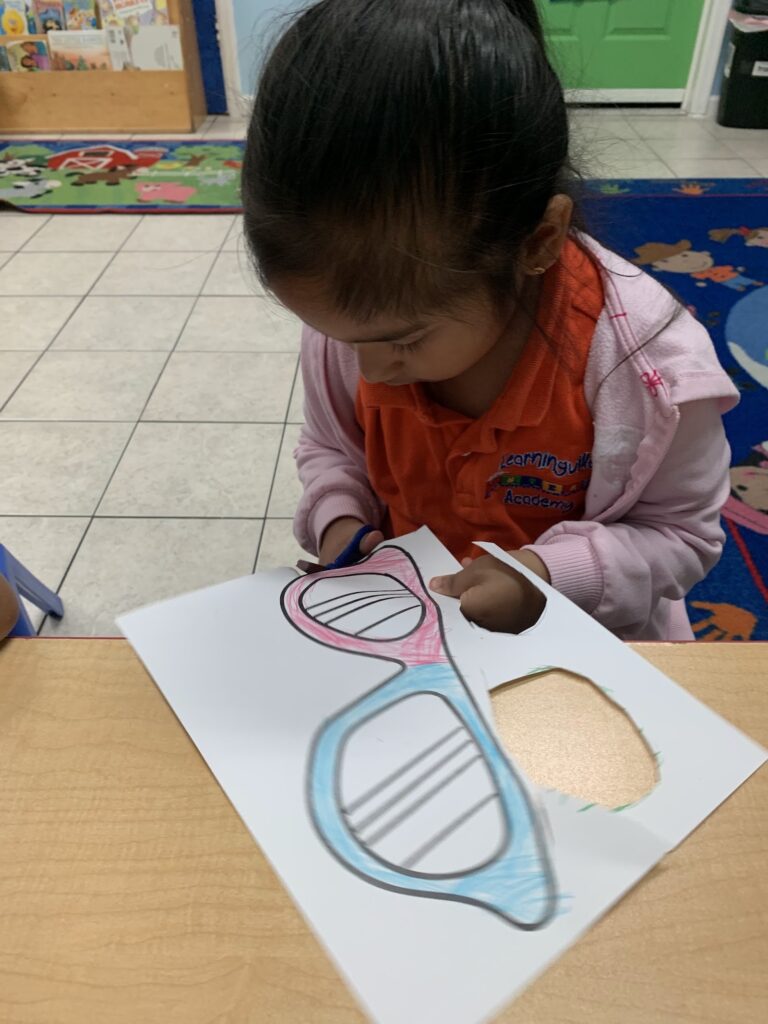 A child coloring and cutting out a paper eyeglasses craft at a classroom table