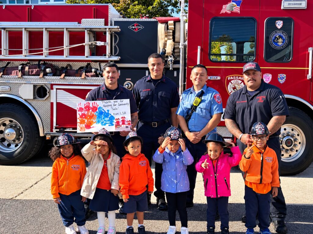 Learningville preschoolers in toy fire helmets with Passaic firefighters beside a fire truck, holding a thank-you sign the children made